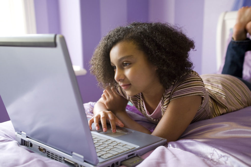 A young girl lays on her stomach on a bed while looking at a laptop
