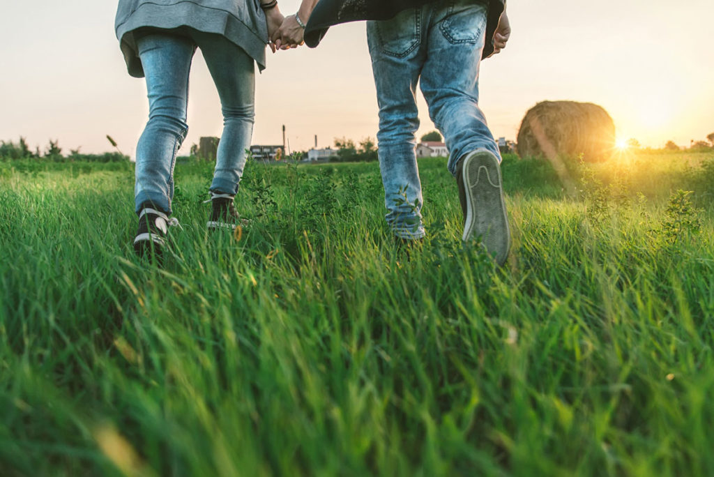 close up of the legs of two teens walking through a green field with a sunset in the background