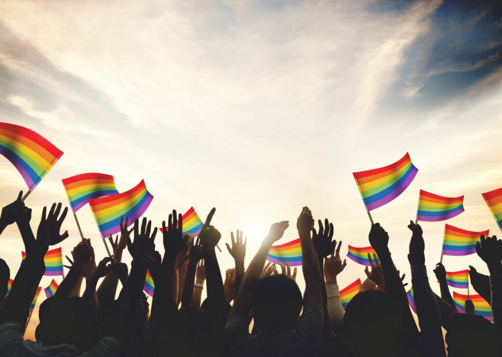a silhouetted group of young people wave rainbow flags for Pride Month